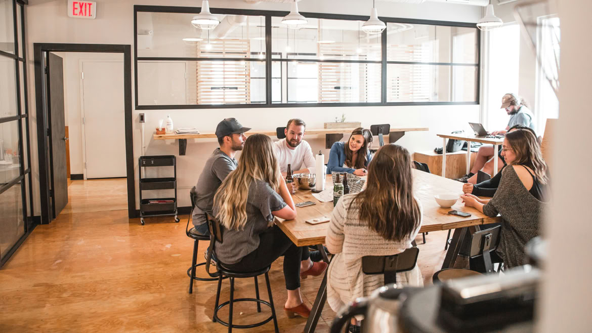A small group is gathered in a bright, creative classroom setting, seated around a table as they discuss ideas. The room resembles an art studio, with colourful materials, sketches and artwork displayed on the walls, giving the meeting an open, imaginative and collaborative atmosphere.